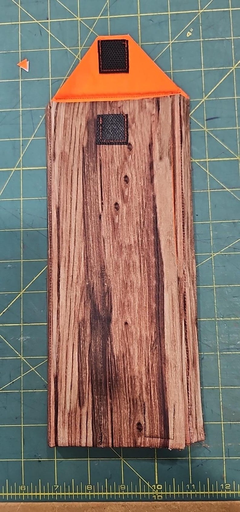 May include: A rectangular phone case with a wood grain pattern. The top is orange with a black square. The case is approximately 25 cm long.