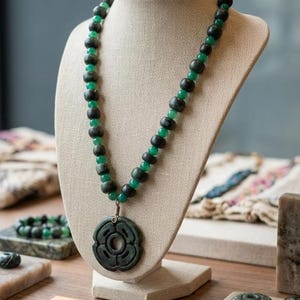 May include: A jade necklace with a large pendant is displayed on a mannequin. The necklace features alternating dark green and black beads. The pendant is a dark green, circular design. Other jade jewelry is visible in the background.