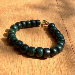 Puede incluir: Una pulsera circular compuesta por numerosas cuentas esféricas, lisas y de color verde oscuro ensartadas. La pulsera cuenta con un pequeño mecanismo de cierre de metal plateado en un extremo, que une los extremos del hilo de cuentas.