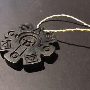 Peut inclure: Un pendentif circulaire gris foncé avec un design géométrique complexe. Le pendentif présente des lignes gravées, des triangles et un motif circulaire central. Une ficelle beige est attachée pour le porter. Le pendentif est probablement en pierre ou en matériau similaire.