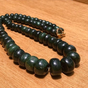 Puede incluir: Un collar de cuentas verde oscuro con un cierre dorado. Las cuentas son redondas y de diferentes tamaños, creando una apariencia texturizada. El collar se muestra sobre una superficie de madera.