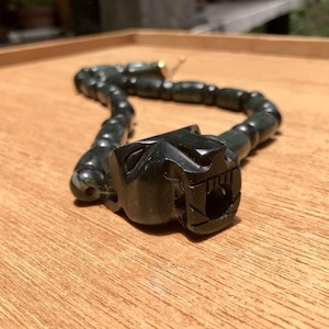 May include: A dark green necklace with a carved panther head pendant. The necklace is made of rounded beads and rests on a wooden surface. The panther head has detailed features.