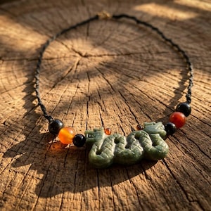 May include: A necklace featuring a carved green stone dragon pendant centred on a black cord. The design includes alternating black and orange-red spherical beads on both sides of the pendant, creating a symmetrical arrangement of colours.