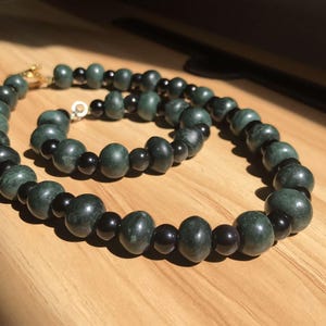 May include: A beaded necklace featuring dark green and black beads. The necklace has a gold clasp and is displayed on a wooden surface. The beads are a mix of rounded and oval shapes, creating a contrasting texture.