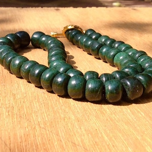 Pode incluir: Um colar de contas verde escuro com fecho dourado, sobre uma superfície de madeira. As contas lisas e arredondadas são exibidas à luz do sol. O colar tem cerca de 45 cm de comprimento.