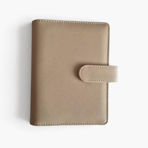 May include: A rectangular beige planner cover with a textured crosshatch finish. The cover features a rounded tab closure on the right side, secured with matching stitching along the edges. The surface has a consistent, fine-grained pattern.