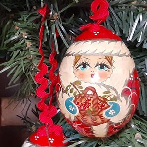 May include: Two hand-painted Christmas ornaments. Each ornament is egg-shaped with a red cap and a red ribbon for hanging. The ornaments feature a painted face, a basket of berries, and red and white decorative patterns.