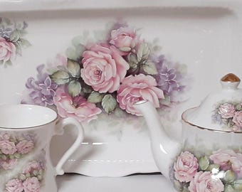 May include: A white porcelain tea set featuring a rectangular serving tray, two mugs, and a teapot, all decorated with painted pink roses, green leaves, and purple accents. Each piece is finished with a delicate gold trim along the edges.