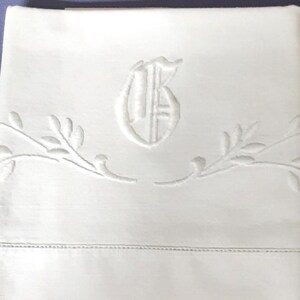 May include: A crisp white rectangular textile featuring a central embroidered monogram of the letter B. The design includes delicate, symmetrical leaf-like vine patterns extending horizontally from the center, with a horizontal hem stitch detail.
