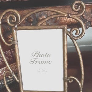 May include: A rectangular photo frame with a hammered gold-tone metal finish and decorative scrollwork accents. The white insert features the text "Photo Frame" and "3.5" x 5" 9cm x 12.5cm" printed in a dark gray serif font.
