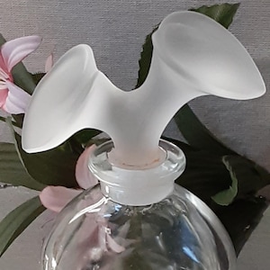May include: A clear glass perfume bottle with a frosted, wing-shaped stopper. The bottle has a rounded base and a short neck. Pink and green floral accents are in the background.