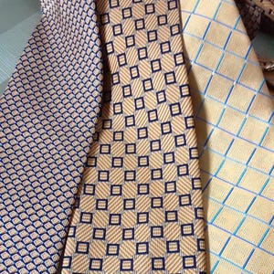 May include: Three patterned neckties in shades of tan and blue. The ties feature geometric designs, including small squares and diamond patterns. The ties are made of a smooth, possibly silk, material.