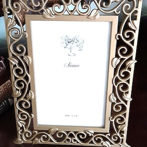 May include: A rectangular gold-colored picture frame featuring an intricate openwork border of swirling vines and leaves. The white insert displays a floral illustration, the name "Sienna" in script, and the printed text "SIZE 4" x 6"."