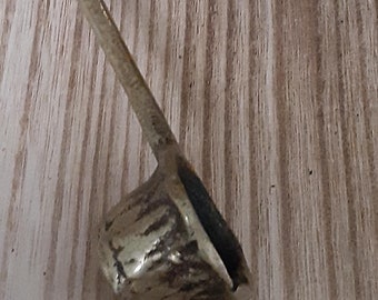 May include: A metallic ladle with a long, slender handle and a small, deep, textured bowl. The surface of the metal has a weathered, uneven finish with dark patina accents, showing a distinct contrast between the smooth handle and the rough bowl.