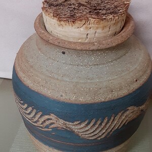 May include: A ceramic jar with a speckled beige finish and a wide, dark blue band around the center featuring a carved, wave-like texture. The jar is topped with a thick, cylindrical cork stopper that has a rough, natural bark-like surface.