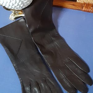 May include: A pair of dark brown leather gloves. The gloves are long, extending past the wrist, and feature stitched details. The gloves are lying on a blue surface.