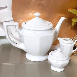 May include: A white ceramic tea set consisting of a large teapot with a lid, a creamer, and a sugar bowl with a lid. Each piece features a faceted, octagonal body design with a subtle embossed decorative border around the rims and lids.