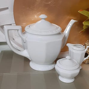 May include: A white porcelain tea set, including a teapot, creamer, and sugar bowl. The teapot has a geometric design and a lid with a small knob. The creamer and sugar bowl have matching details. The set is on a striped surface.