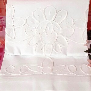 May include: A folded white textile featuring intricate white embroidery. The fabric displays a large, central floral motif with rounded petals and a secondary border pattern of smaller, stylized flower shapes along the bottom edge.