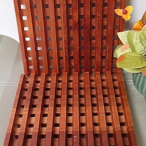 May include: Two wooden trivets with a grid pattern. The trivets are made of reddish-brown wood and have a series of vertical and horizontal slats, creating a square grid design. The trivets are designed to protect surfaces from hot dishes.