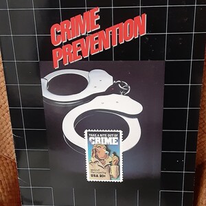 May include: A black and white Crime Prevention poster featuring a pair of handcuffs and a postage stamp. The stamp shows McGruff the Crime Dog and the text "Take a Bite Out of Crime". The title "CRIME PREVENTION" is in red.