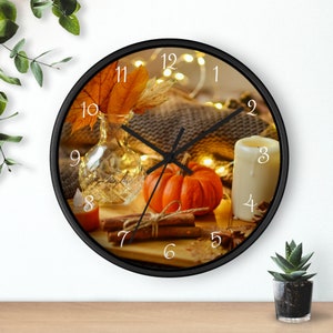 Puede incluir: Un reloj redondo negro con un diseño de otoño. La esfera del reloj presenta una calabaza, palitos de canela, una vela y hojas de otoño. Los números en la esfera del reloj son blancos.