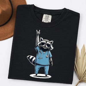 May include: Black t-shirt featuring a cartoon raccoon wearing sunglasses and blue scrubs, giving a rock-and-roll hand gesture. The raccoon has a stethoscope around its neck. The shirt has a Comfort Colors tag.