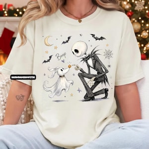 May include: A cream-colored t-shirt featuring a graphic of a skeleton character in a pinstripe suit kneeling to touch the nose of a ghost dog. The design includes the text "Boop," a crescent moon, bats, stars, and a spider web in black and orange.