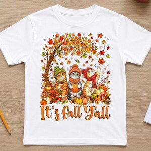 May include: A white short-sleeve t-shirt featuring a graphic of three cats wearing knit hats and scarves under an autumn tree with falling leaves, pumpkins, and mushrooms. The text "It's fall Y'all" is printed in orange below the illustration.