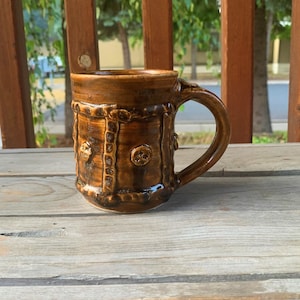May include: A brown ceramic mug with a textured design and a handle. The mug has a rustic look and is decorated with raised circles.