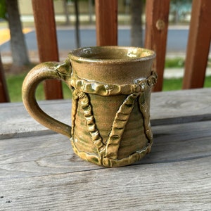 May include: A handmade ceramic mug with a green and brown glaze. The mug has a unique design with a braided rope pattern around the body.