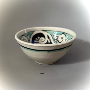 May include: A small white ceramic bowl with a blue and black floral design. The bowl has a black and green rim.