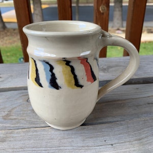 May include: A white ceramic mug with a colorful striped design. The stripes are black, yellow, blue, and red.