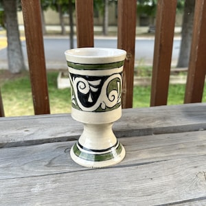 May include: A white ceramic cup with a green and black geometric design. The cup has a pedestal base.
