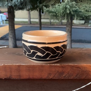 May include: A small, cream-colored ceramic bowl with a black braided design around the rim.