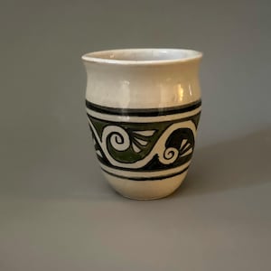 May include: A white ceramic mug with a green and black geometric design. The design features a repeating pattern of swirls and leaves.