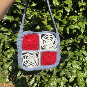 May include: A crocheted bag with a blue background and four squares. The squares are red, white, and black. The bag has a long strap.