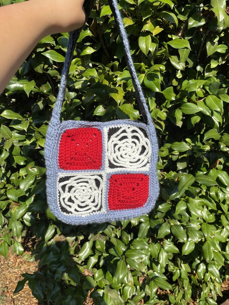 May include: A crocheted bag with a blue background and four squares. The squares are red, white, and black. The bag has a long strap.