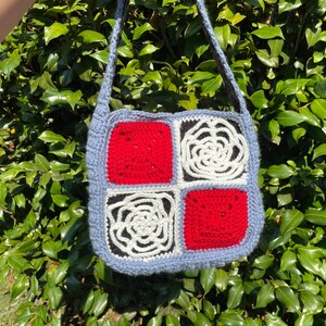 May include: A crocheted bag with a blue, red, white and black pattern. The bag is square shaped and has a long strap.