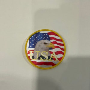 May include: A round, white patch with a yellow border. The patch features a bald eagle with a red, white and blue American flag in the background. The word "USA" is written in gold letters below the eagle.