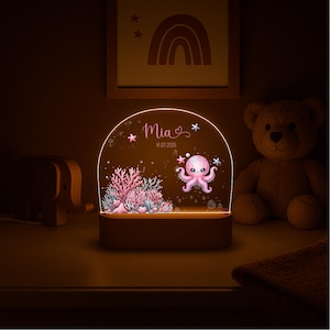 Peut inclure: Veilleuse personnalisée avec un motif de poulpe, le nom "Mia" et la date 14.07.2025. La lumière a une base en bois et un panneau en acrylique transparent avec des éléments roses et coralliens. Un ours en peluche et un éléphant en bois sont en arrière-plan.