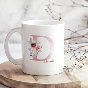 Op de afbeelding: Witte keramische mok met een grote roze letter "D" versierd met bloemmotieven in roze, bordeauxrood en groen. De naam "Daniela" staat eronder gedrukt. De mok staat op een houten dienblad.