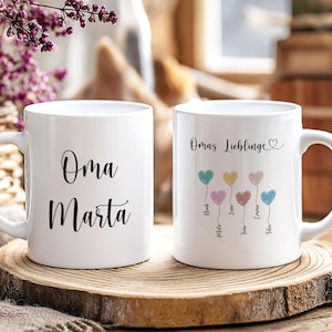 Könnte beinhalten: Zwei weiße Keramikbecher mit schwarzer Schrift. Auf einem Becher steht "Oma Marta", auf dem anderen "Omas Lieblinge" mit herzförmigen Ballons und Namen. Die Becher stehen auf einer Holzscheibe.