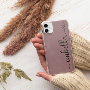 Può includere: Una custodia trasparente per telefono con retro color malva, con il nome "Isabella" in elegante scrittura nera. Un piccolo cuore è disegnato sopra il nome. La custodia è tenuta tra le mani di una persona, con uno sfondo neutro.