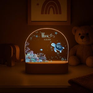 K&ouml;nnte beinhalten: Ein halbkreisf&ouml;rmiges Acryl-Nachtlicht mit einer farbenfrohen Unterwasserszene, die eine Meeresschildkr&ouml;te, ein Korallenriff und Blasen zeigt. Das Licht ist mit dem Namen Theo und dem Datum 16.05.2024 auf einem Holzsockel personalisiert.