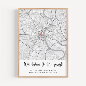 Op de afbeelding: Een zwart-witte kaart van Keulen, Duitsland, met een rode hartpin die een locatie markeert. De tekst onder de kaart luidt "Wir haben Ja gesagt", wat in het Nederlands "We hebben ja gezegd" betekent. De datum "23. Juli 2023" en de namen "Nina & Marco" zijn ook opgenomen. De coördinaten "Köln 50° 56'0.6"N 6° 56'43.9"E" staan onder de namen.