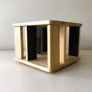 May include: A light-colored wooden cube-shaped storage unit with vertical supports and black inserts. The unit has a rounded top and base, and is designed to hold items. The overall design is simple and modern.