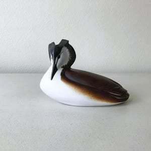 May include: A ceramic figurine of a bird, featuring a white body with a brown back and a black head. The bird is in a resting pose, with its head slightly lowered. The figurine is set against a plain white background.