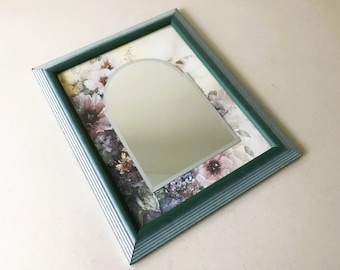May include: A rectangular framed mirror with a green and white striped border. The mirror has an arched top and is set against a floral print background with pink and purple flowers.