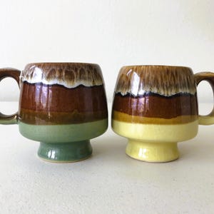 May include: Two ceramic mugs with brown, green, and yellow glaze. The mugs have a wide base and a rounded shape.